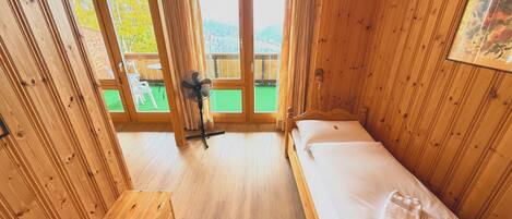 A wooden cabin room with a bed, a fan, and a view of a green outdoor area.