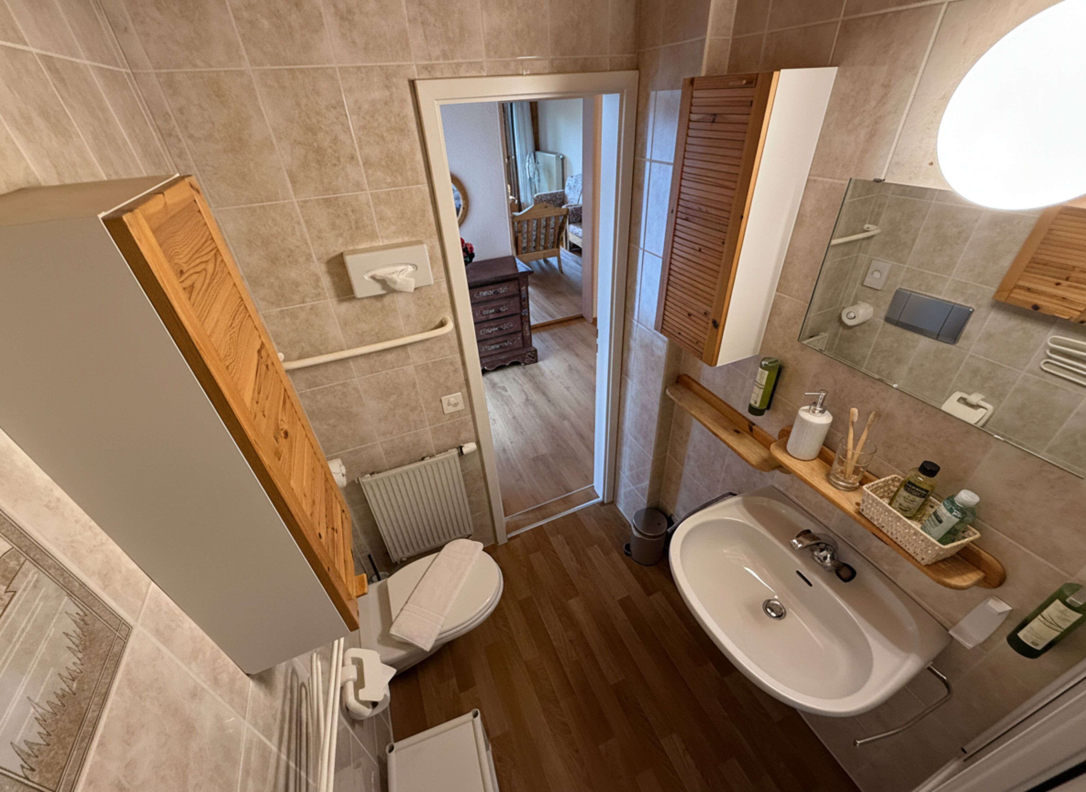 A modern bathroom with a toilet, sink, and shower area. There is a wooden door leading to a hallway with a chair and a radiator.