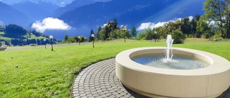 Una fuente circular con agua brotando del centro, rodeada por una zona pavimentada donde se aprecia una tapa de alcantarilla. Al fondo, montañas y un cielo despejado.