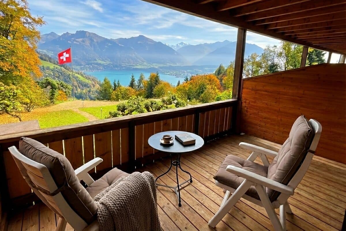 Terraza de madera con dos sillas y una mesita, con vistas a un paisaje montañoso y un lago, e izada una bandera.