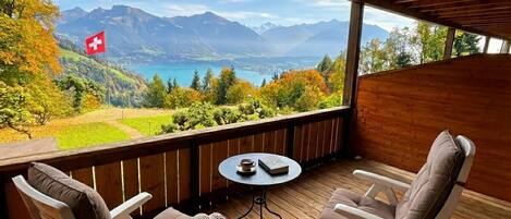 A wooden deck with two chairs and a small table, overlooking a mountainous landscape with a lake and a flag.