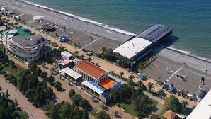 An aerial view of a coastal area with a beach, a large building with a flat roof, and a clear blue sky with scattered clouds. - Beautiful Studio Apartment in Batumi (Batumi)