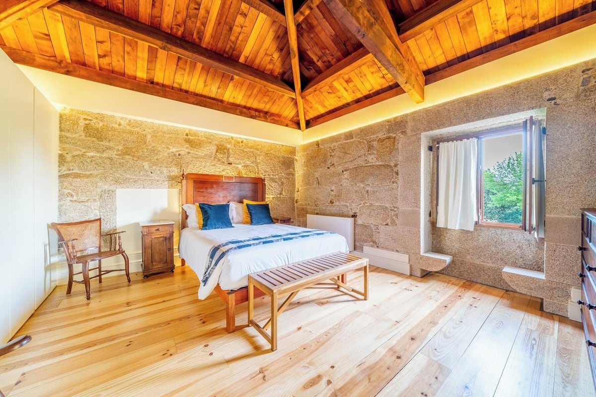A bedroom with a stone wall, wooden ceiling beams, a bed with pillows, a bench, and a window with a view.