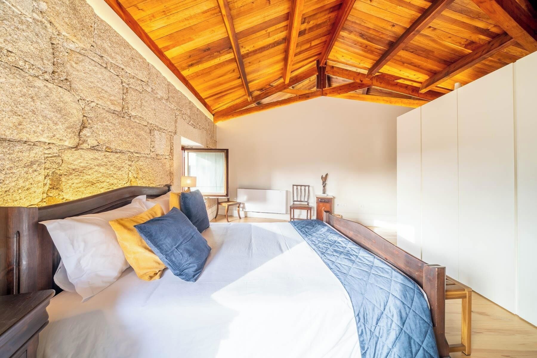 Un dormitorio con pared de piedra, vigas de madera en el techo, una cama con ropa de cama blanca y azul, una mesita de noche de madera y una silla.