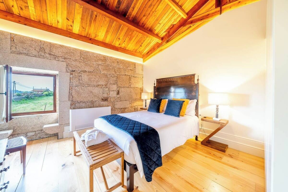 A bedroom with a stone wall, wooden ceiling, a bed with a headboard, bedside tables, and a bench.