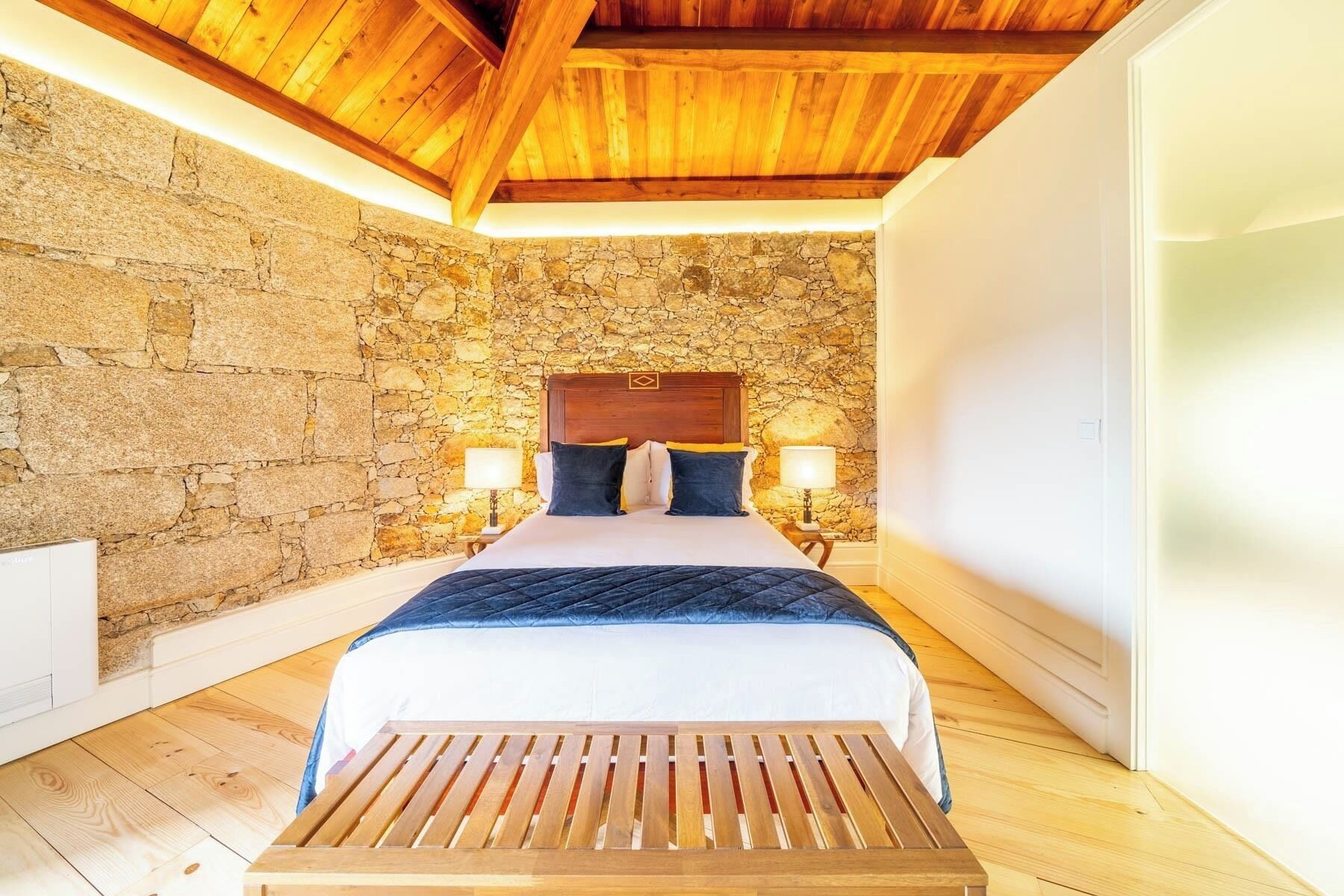 Quarto com parede de pedra, teto de madeira, cama com cabeceira de madeira, duas luminárias de cabeceira e um banco de madeira.