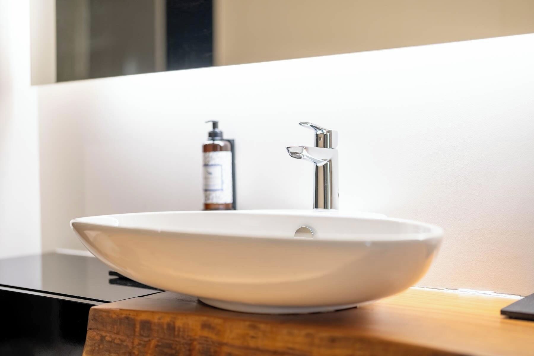 A white vessel sink with a modern faucet, a bottle of soap, and a wooden surface.