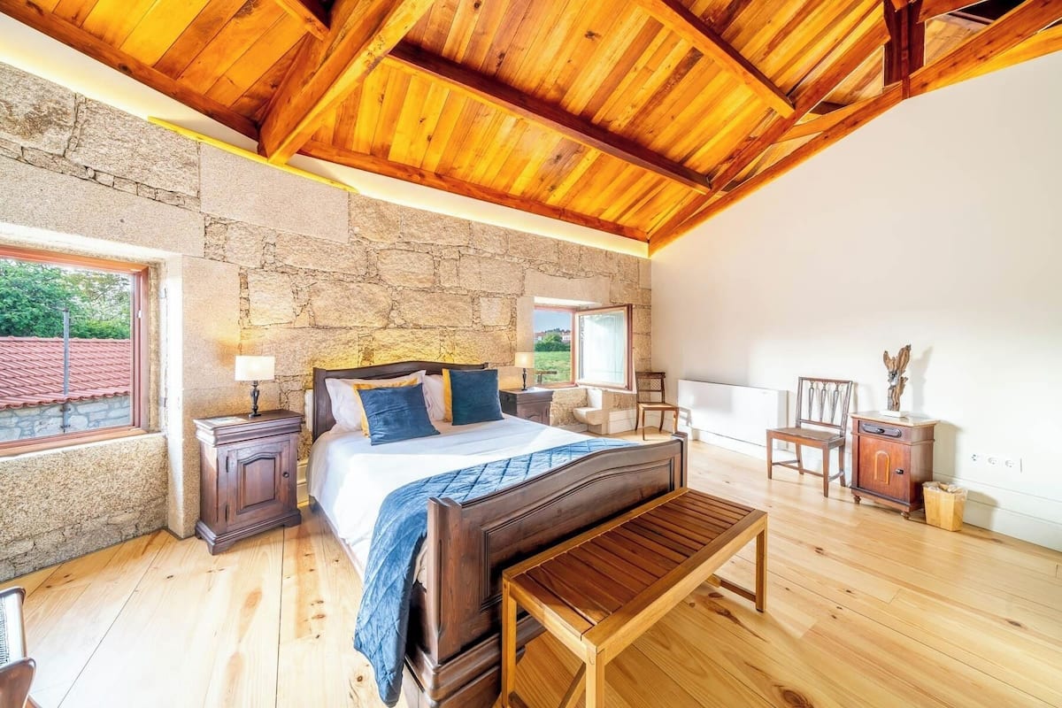 A bedroom with a stone wall, wooden ceiling, a bed with pillows, a nightstand, a wooden bench, and a chair.