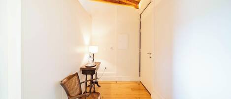 Un pasillo estrecho con piso de madera, una silla, una mesita con lámpara y una puerta.