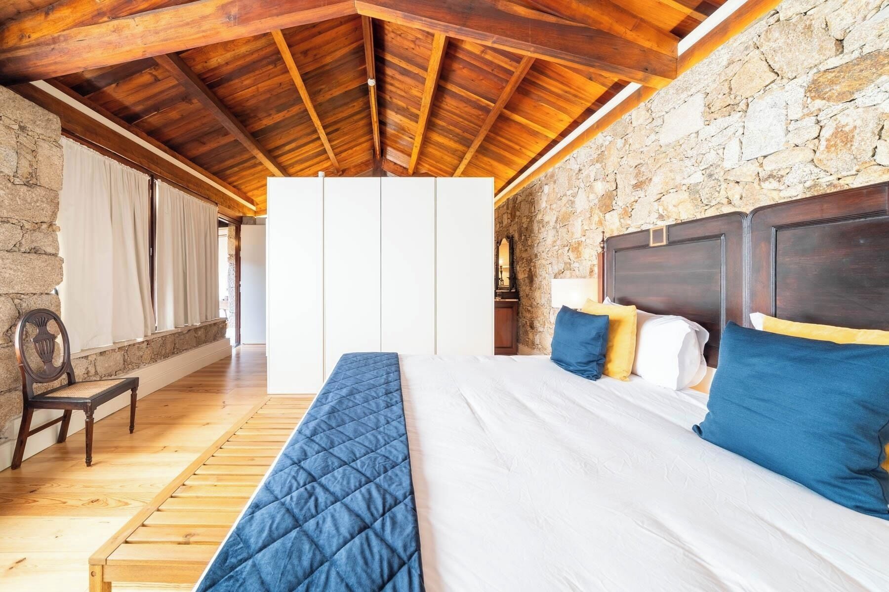 A bedroom with a stone wall, wooden ceiling beams, a bed with blue and white bedding, and a bench.