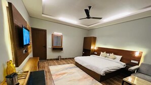 A modern hotel room with a bed, a desk, a chair, a TV, and a ceiling fan. - The Foresta (Dhari)