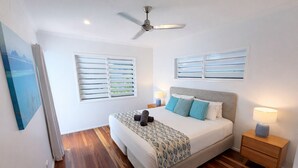 Un dormitorio con cama, dos mesitas de noche, ventilador de techo y dos ventanas con persianas.