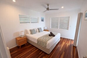 Une chambre à coucher comprenant un lit, des tables de chevet, un ventilateur de plafond et deux fenêtres équipées de volets.