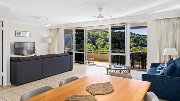 A modern living room with a sofa, a TV, and a dining table. There is a balcony with a view of the forest. - Poinciana 109 & Buggy- Hamilton Island (Whitsundays)