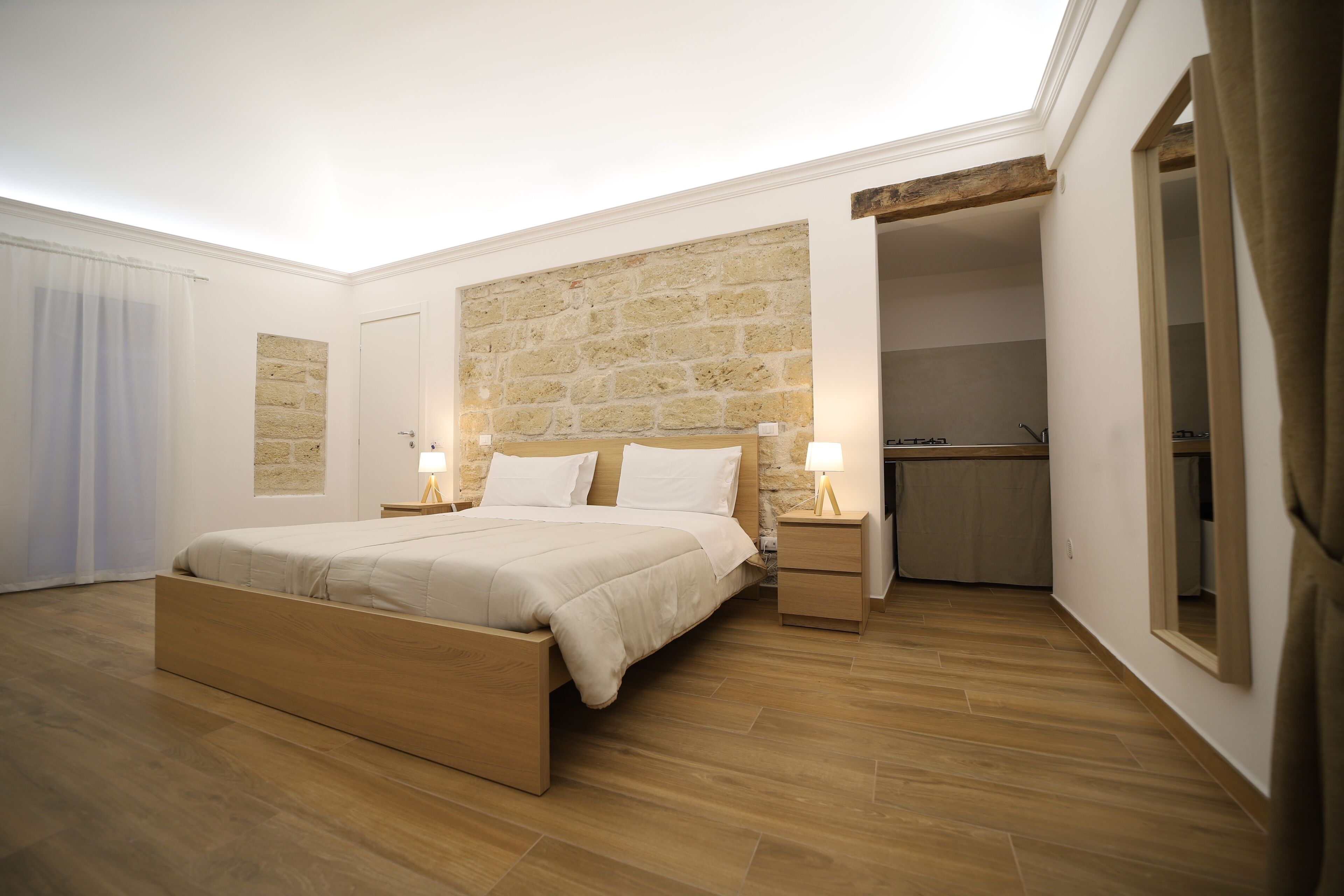 A modern bedroom with a wooden bed, stone wall, and a small kitchen area.