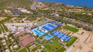 Eine Vogelperspektive zeigt einen luxuriösen Urlaubsort mit mehreren Schwimmbecken, Tennisplätzen und einem Strandbereich.