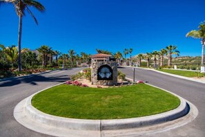 A well-maintained road with a stone monument featuring a decorative emblem, surrounded by a circular grassy area and palm trees.