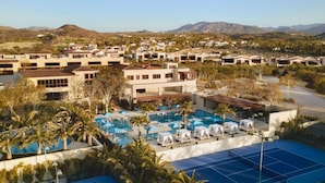 Un resort con varias edificaciones, un área de piscina con sillones de descanso y una cancha de tenis.