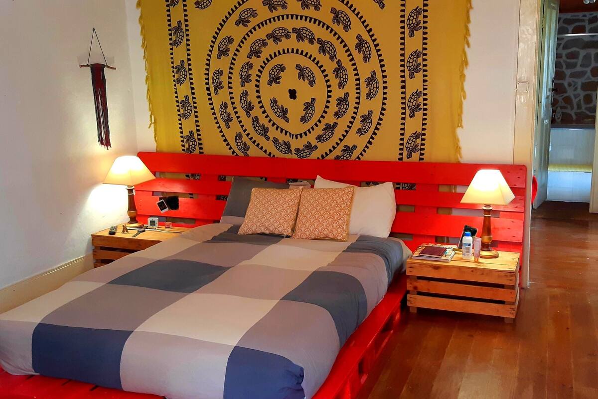 Una cama con colcha a cuadros, dos mesitas de noche con lámparas, un cabecero rojo y un tapiz con estampado de tortuga en la pared.