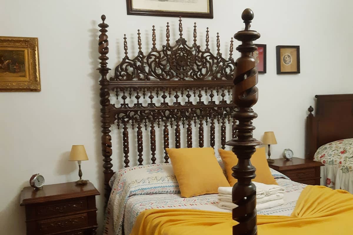 Una cama con dosel, un cabecero de madera, una mesita de noche con lámpara y una alfombra estampada en el suelo.