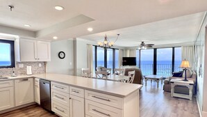 A modern kitchen with a large island, dining area with a view, and a living room with a TV and balcony. - 2BR/2BA End Unit, Pool , Prvt Covered Parking, Seasonal Beach Serv. (Panama City Beach)
