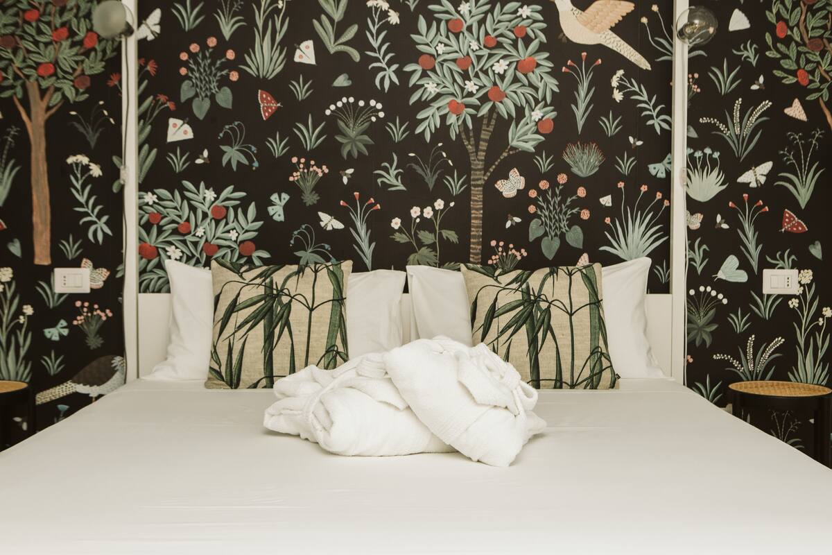 A bed with white linens and pillows, a dark floral wallpaper, and a small round table with a lamp.
