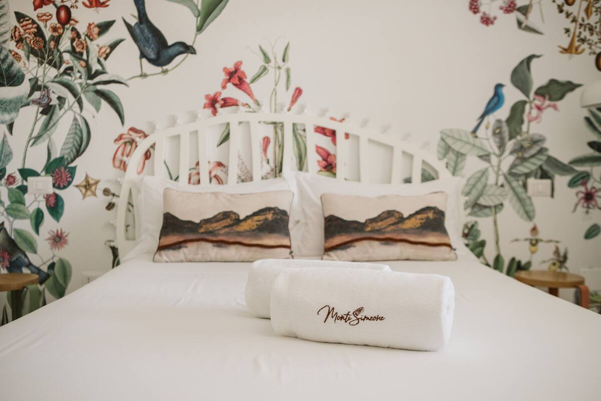 A bed with white bedding, two pillows with a mountain landscape design, and a folded white towel with the text "Montagne".