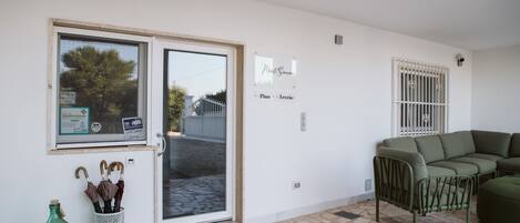 Una habitación con un sofá verde, una pared blanca con un cartel, una puerta con un panel de vidrio y un suelo de baldosas.
