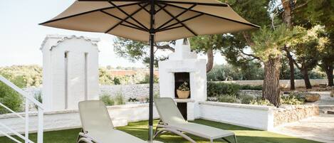 Two lounge chairs under a large umbrella in a landscaped garden area with white walls and a stone fireplace.