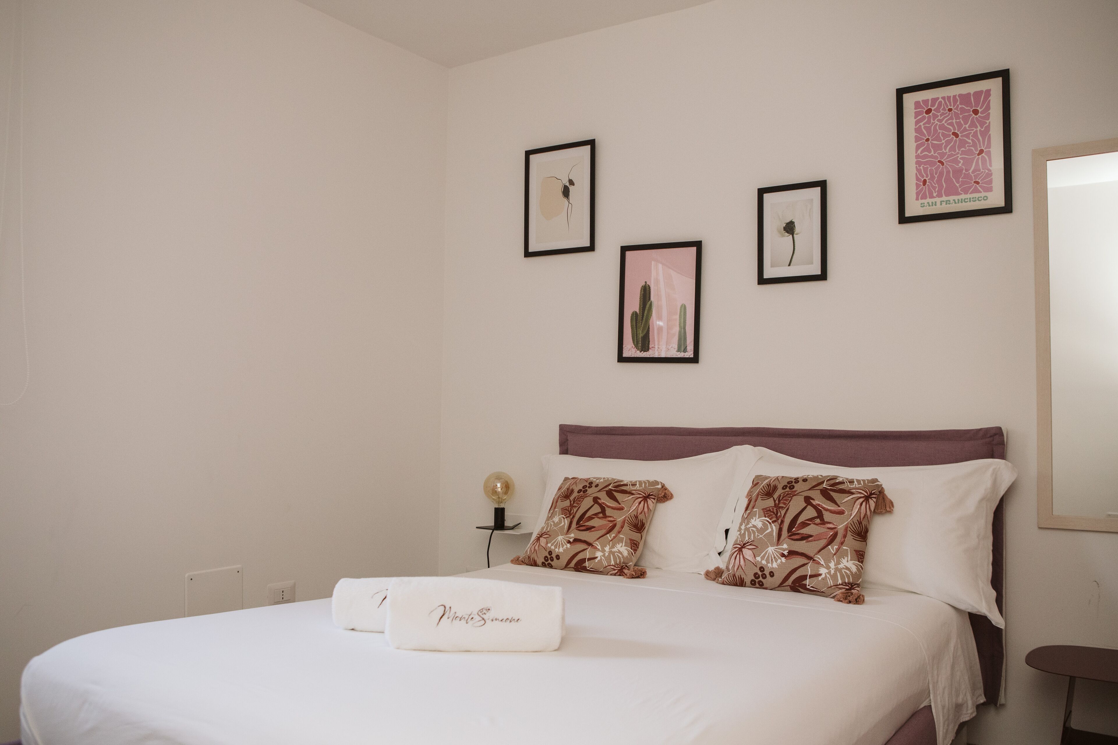 A neatly made bed with a white comforter and decorative pillows, a bedside table with a lamp, and framed artwork on the wall.