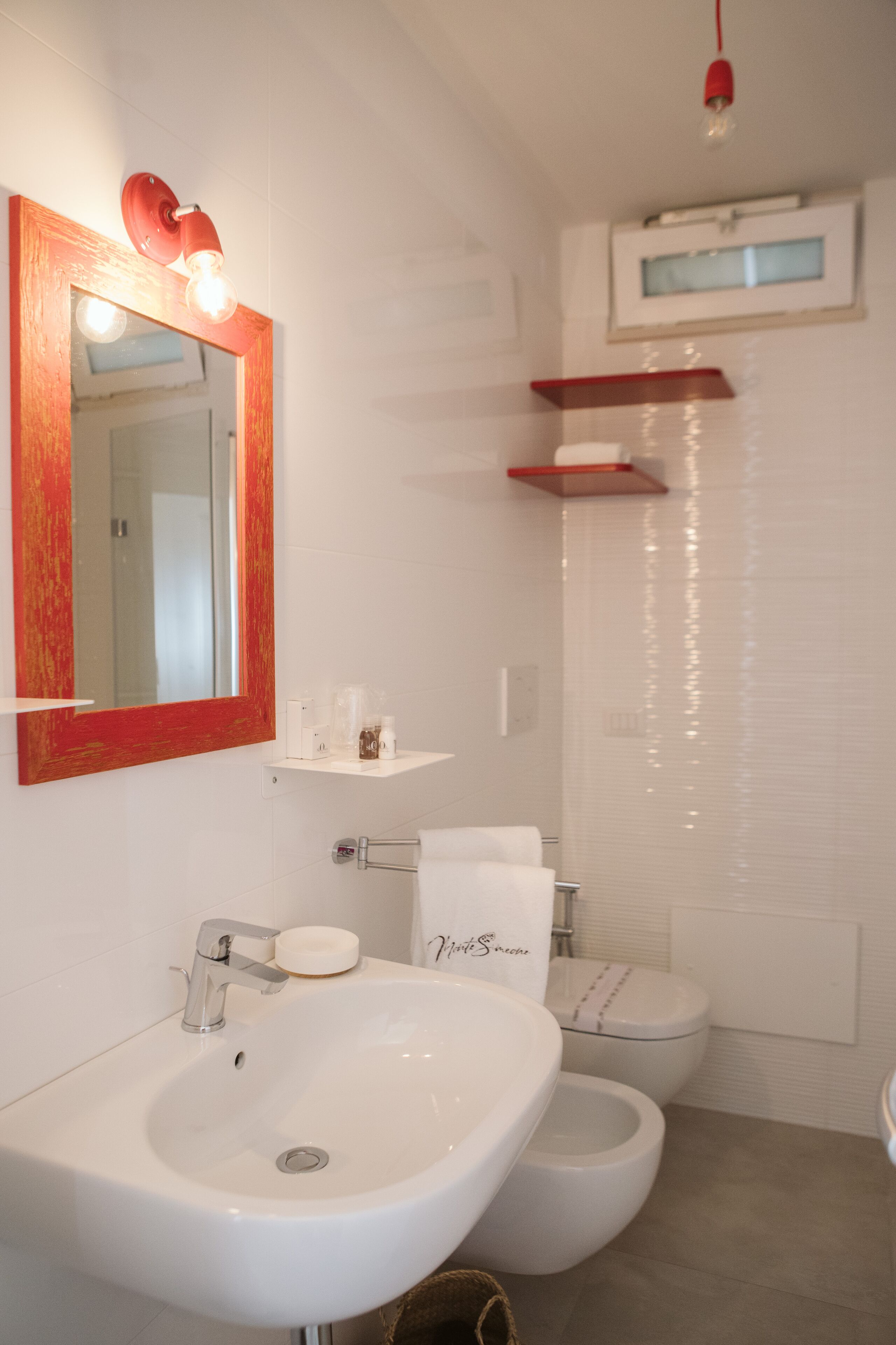 Une salle de bain avec un lavabo blanc, un miroir avec un cadre rouge, des toilettes et une étagère contenant des articles de toilette.
