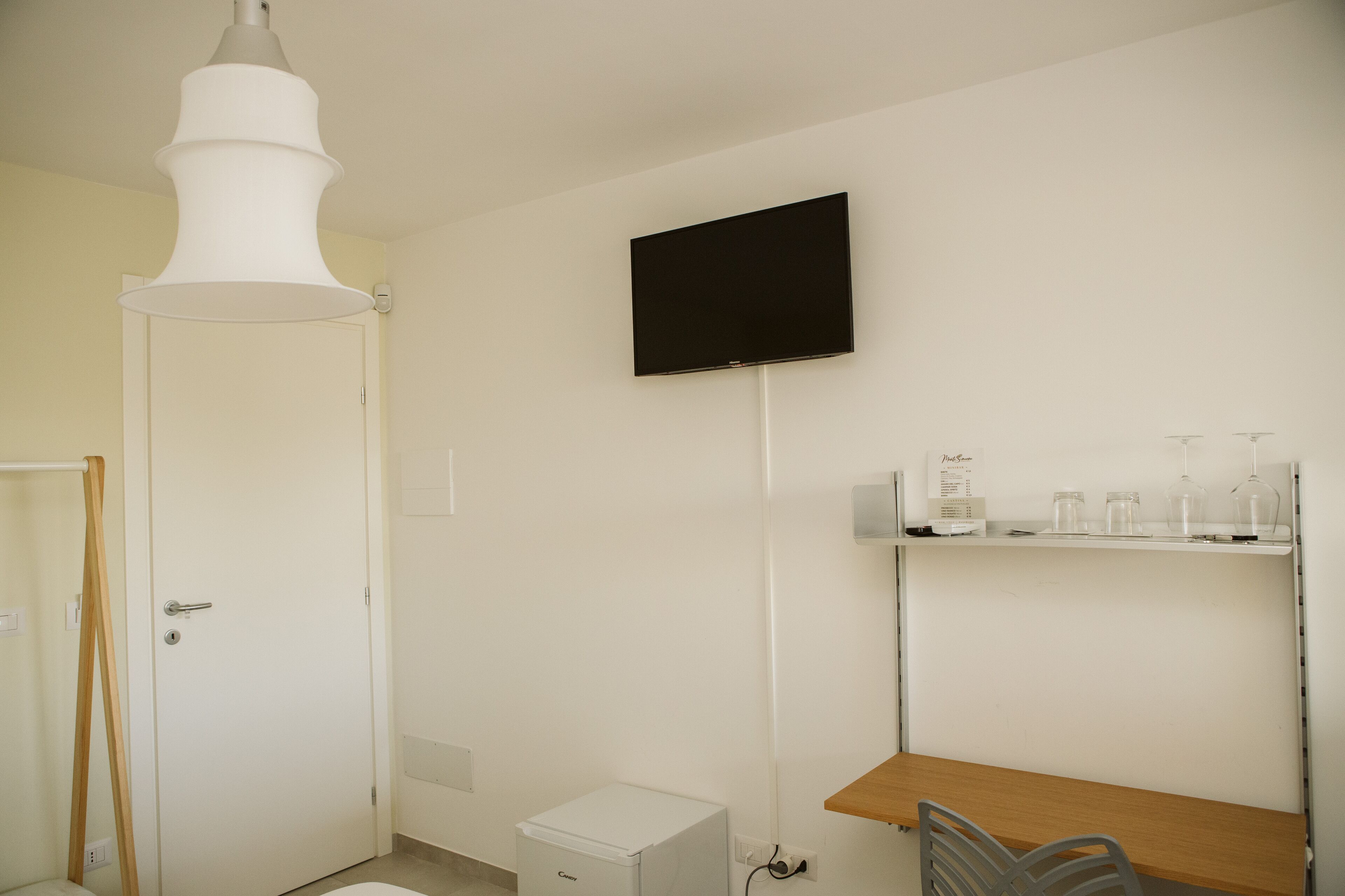 Ein Zimmer mit weißer Tür, einem kleinen, weißen Kühlschrank, einem Holztisch, einem Stuhl, einem Wand-Fernseher und einem Regal mit Glasbehältern und einer Flasche.