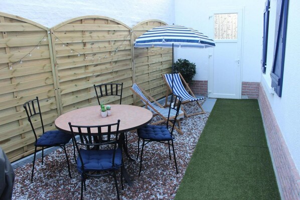 A patio with a round table, four chairs, a striped umbrella, and a small garden area with artificial grass.