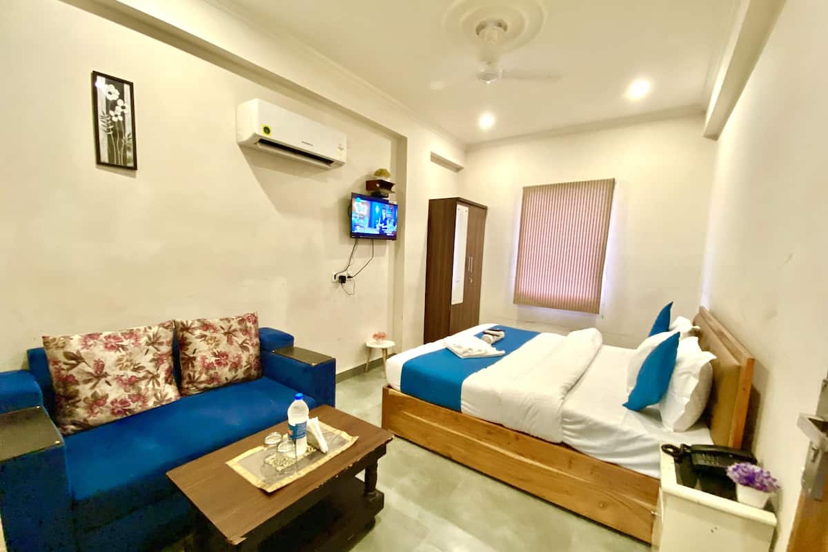 A hotel room with a blue sofa, a wooden bed, a small table, an air conditioning unit, a television, and a window with blinds.
