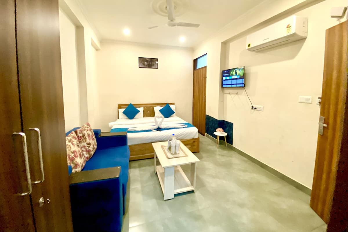 A hotel room with a bed, a blue sofa, a small table, a ceiling fan, an air conditioning unit, and a TV mounted on the wall.