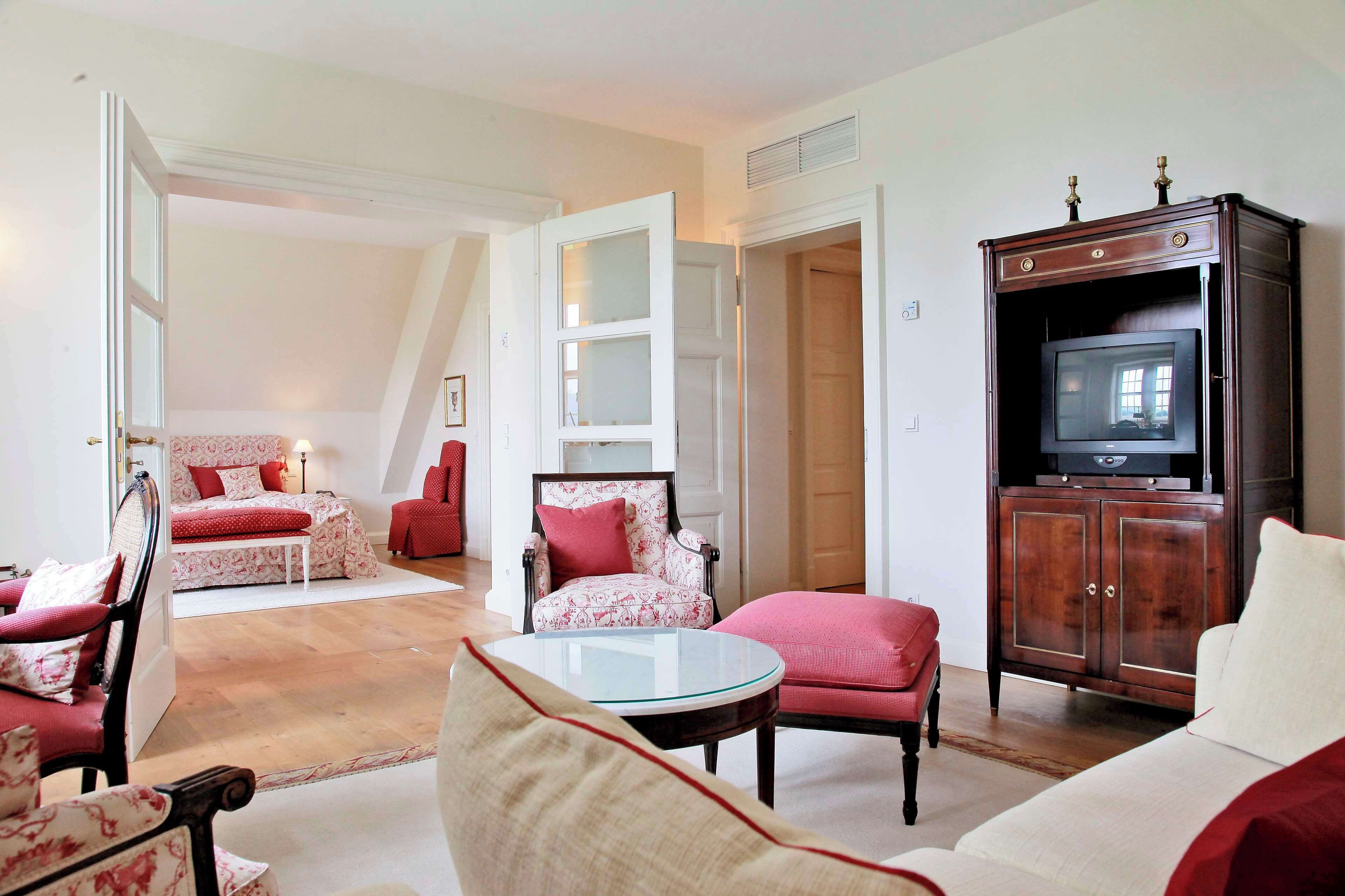 A spacious living room with a large television on a wooden cabinet, a red sofa, a patterned armchair, and a glass coffee table.