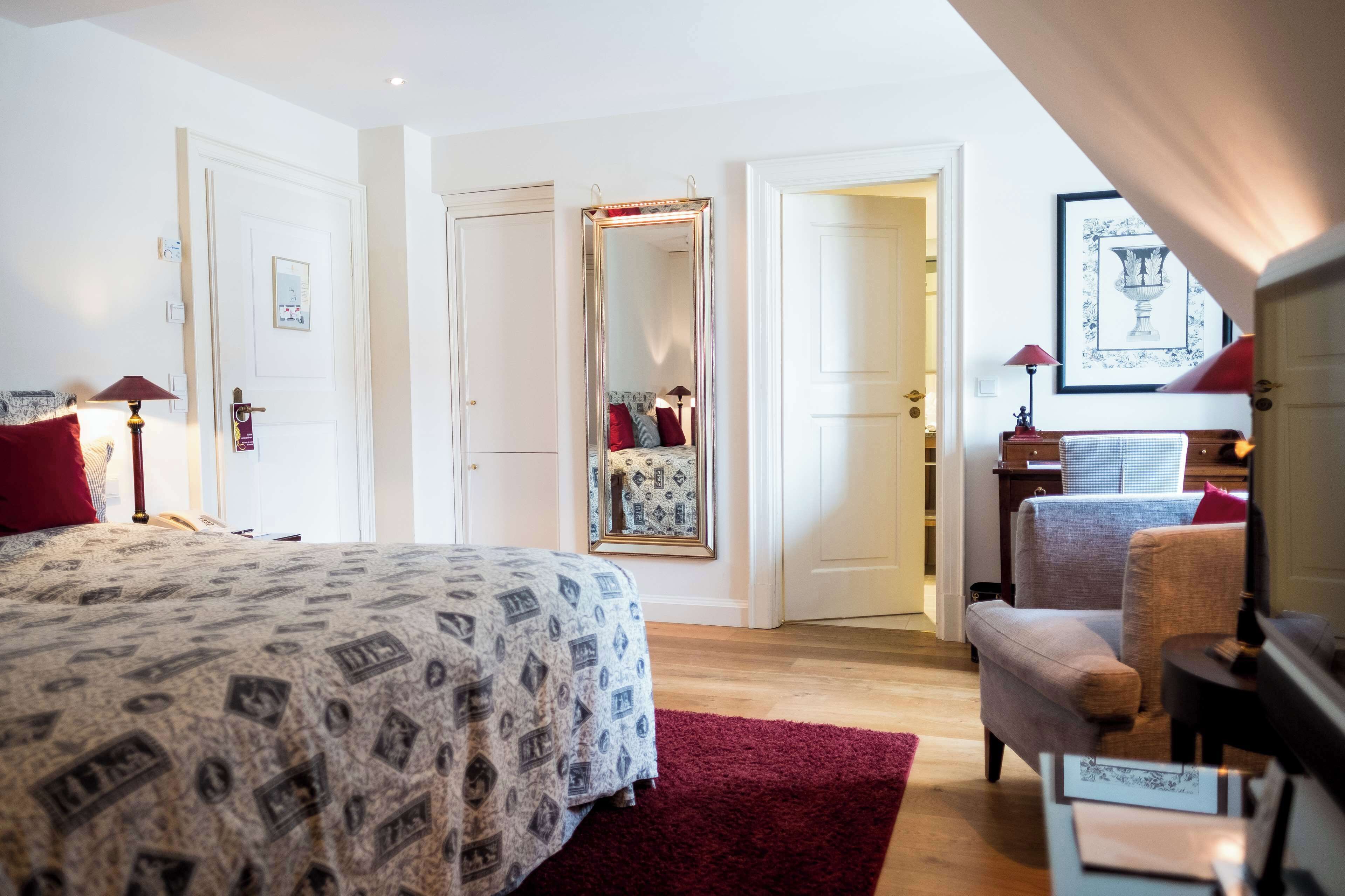 Habitación de hotel con una cama grande, un rincón de descanso con una silla, un espejo y un cuadro en la pared.