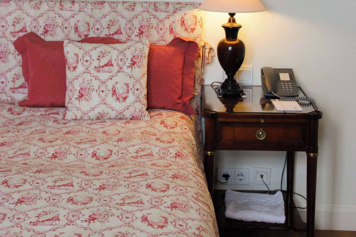 Een keurig opgemaakt bed met bloemenbedrukking, een bedlampje, een telefoon en een lijstje aan de muur.