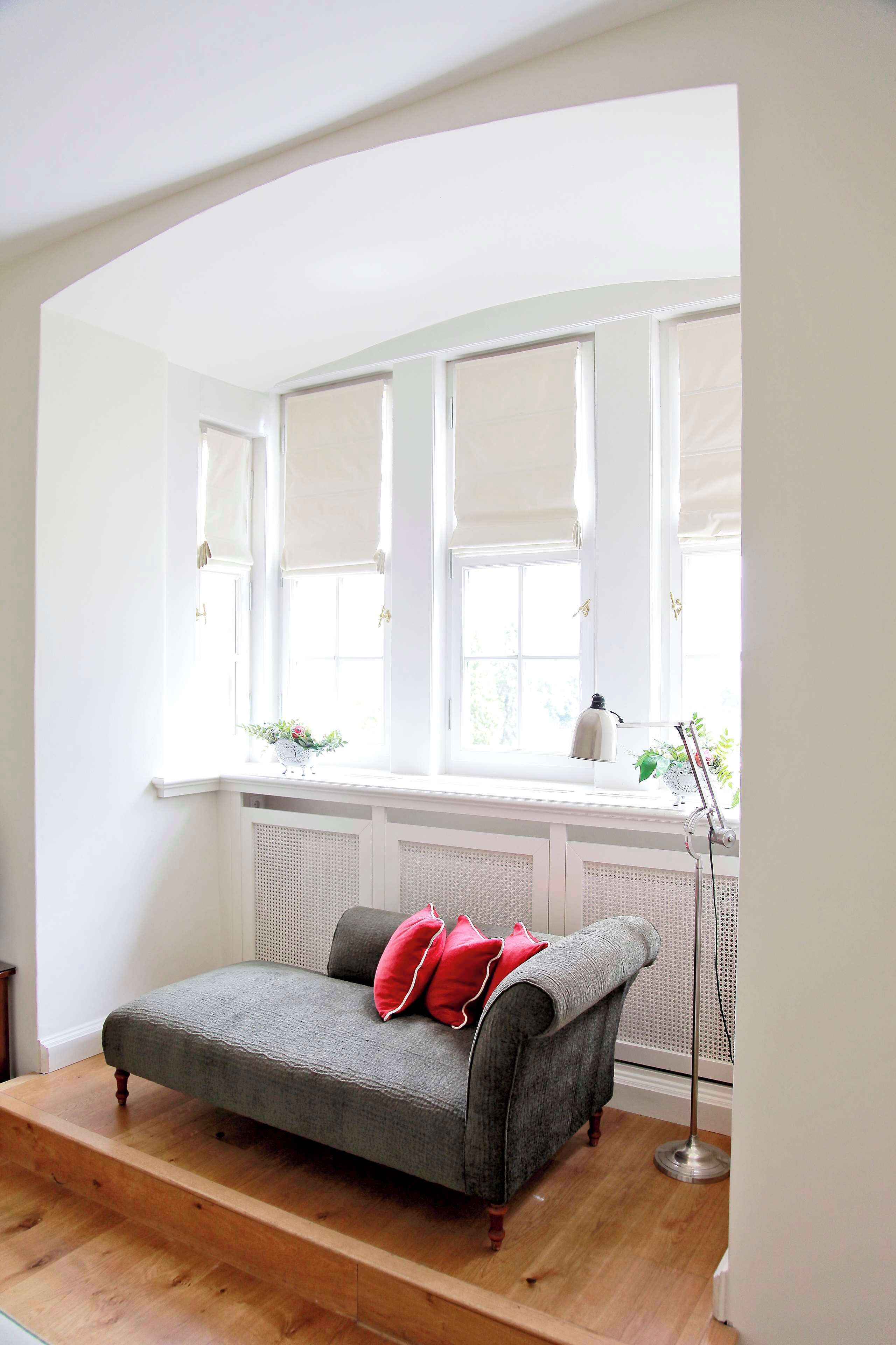 A cozy room with a grey sofa, red cushions, a floor lamp, and a window with white blinds.