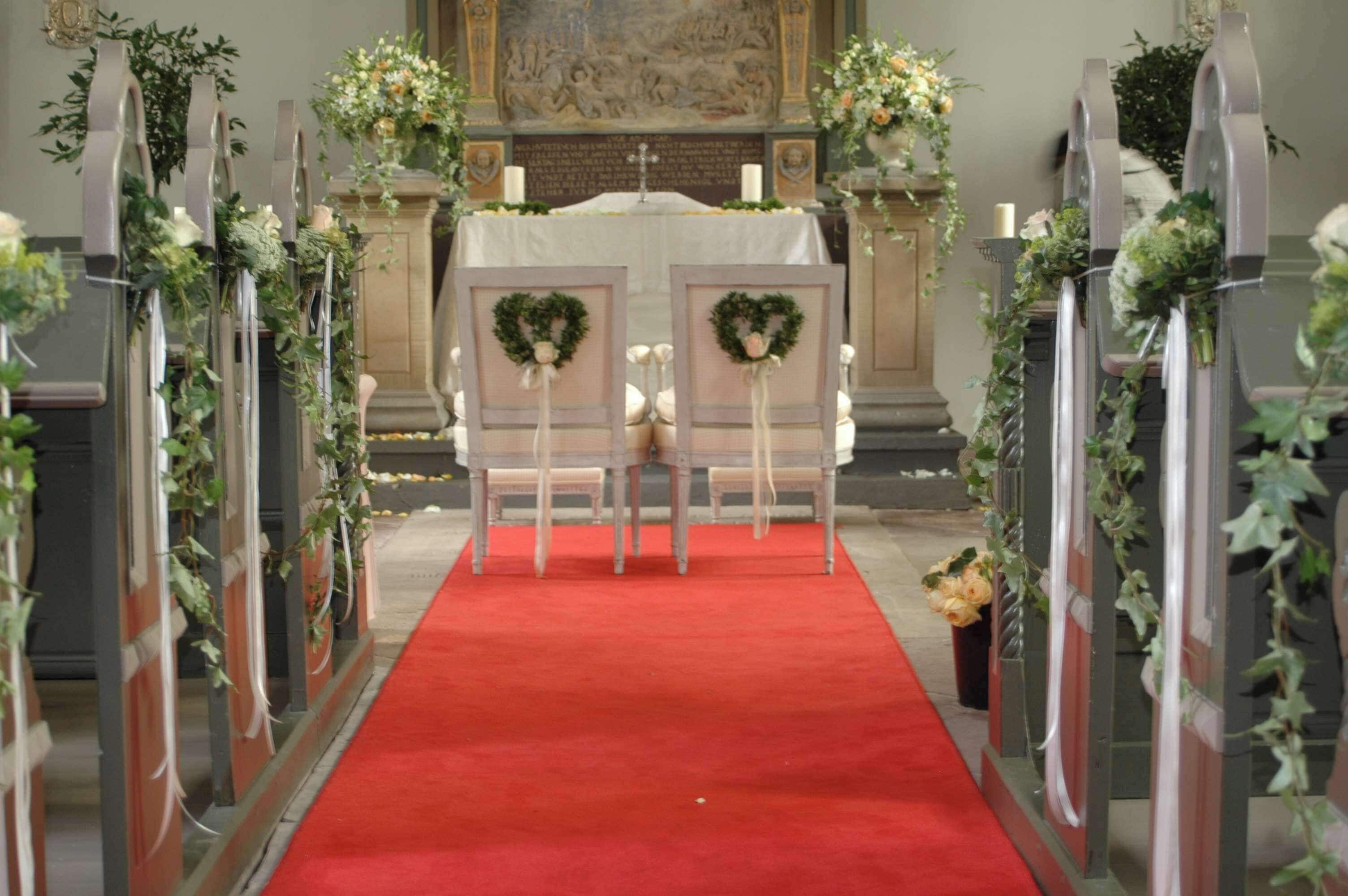 Ein Kirchengang mit Kirchenbänken, geschmückt mit Grünpflanzen und Blumen, der zu einem Altar mit rotem Teppich führt.