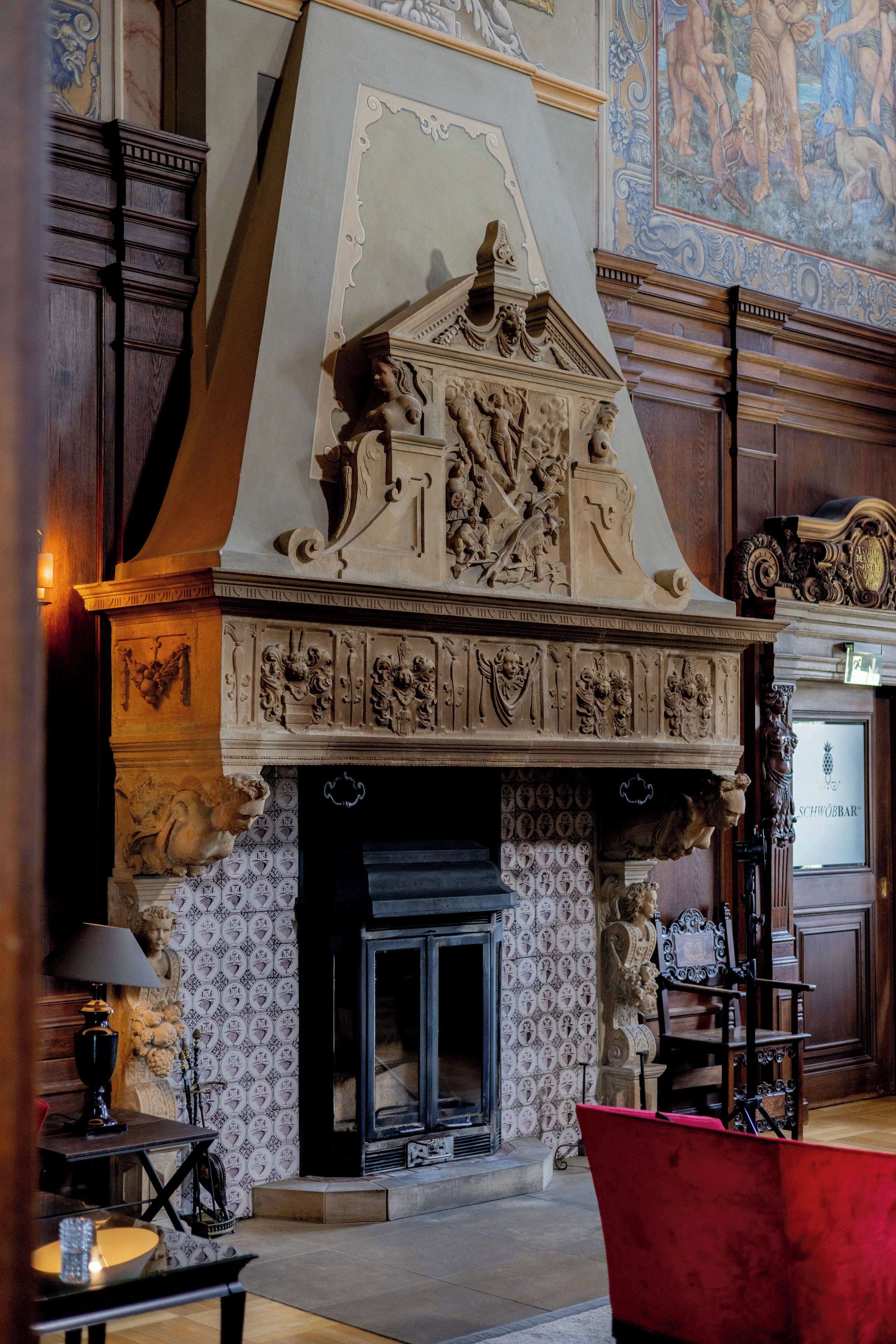 A fireplace with intricate carvings and a mantelpiece, a patterned wallpaper, and a red armchair in a room with wooden paneling and a large mural on the wall.