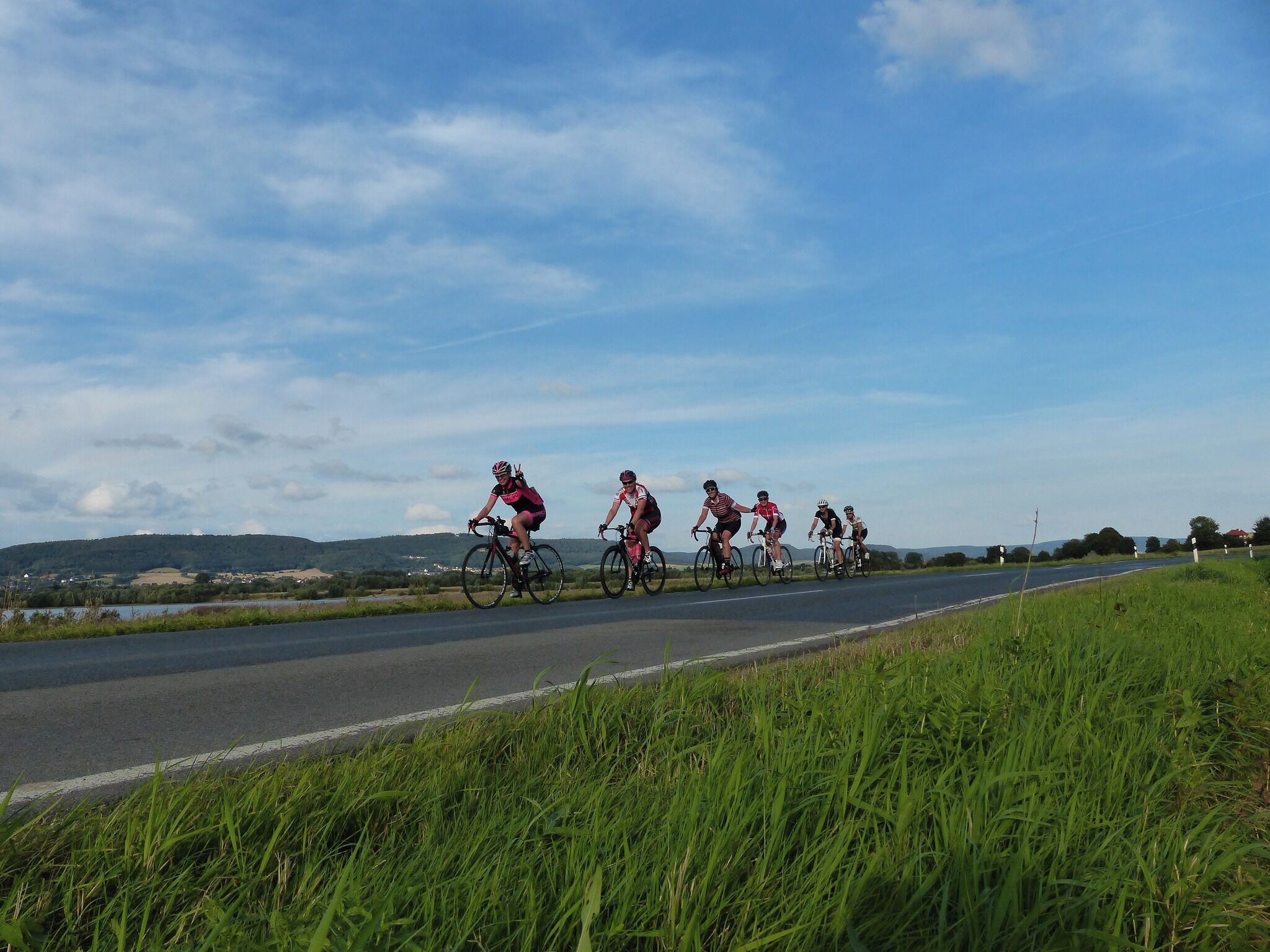 Eine Gruppe Radfahrer auf einer Straße mit grünen Rasenflächen am Rand und einer hügeligen Landschaft im Hintergrund.