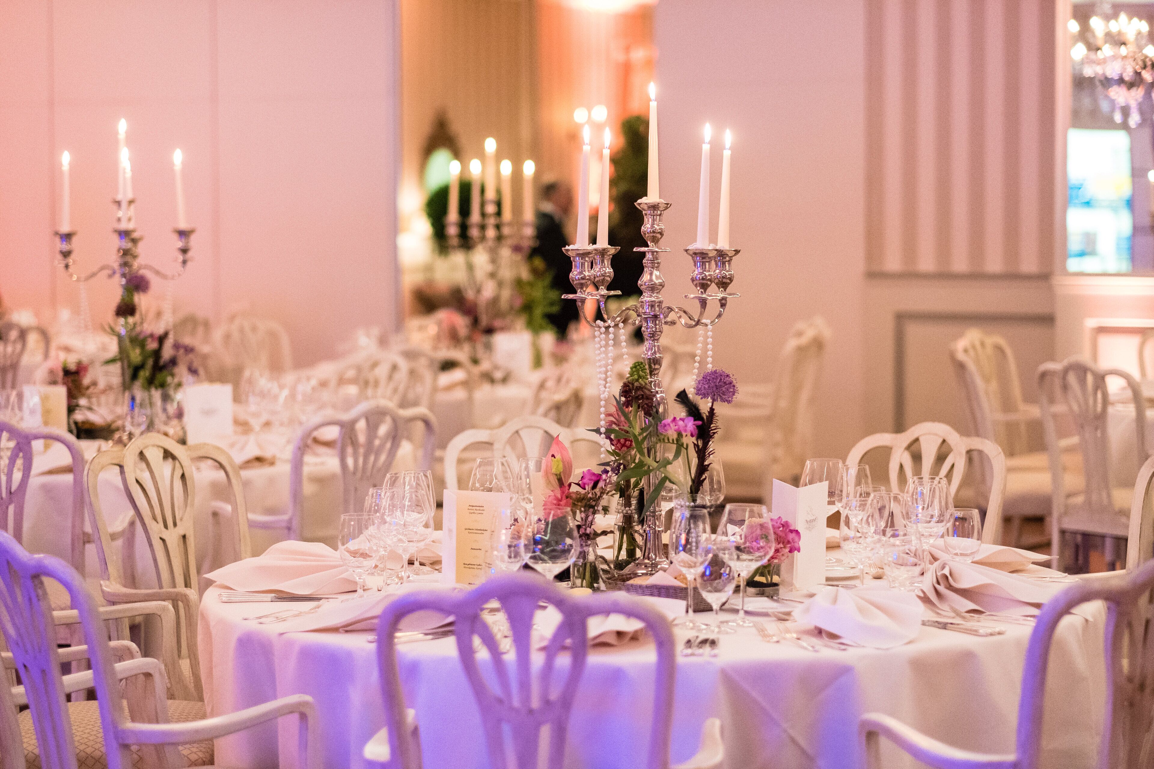 Ein Bankettsaal mit gedeckten Tischen für eine festliche Veranstaltung, mit weißen Tischdecken, Kerzenständern und floralen Tischgestecken.