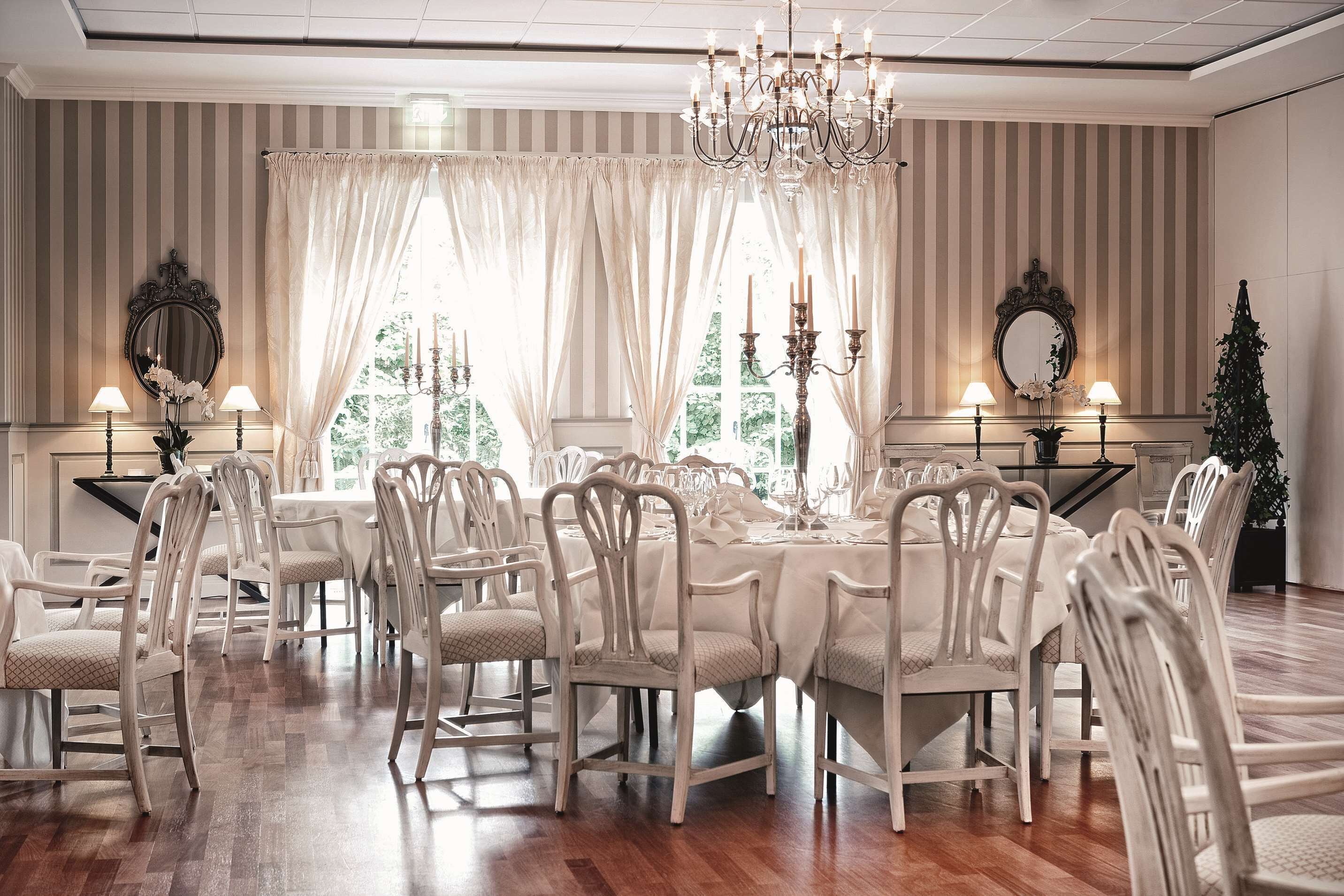 A dining room with a large table set for a meal, surrounded by white chairs, with chandeliers and wall-mounted mirrors.