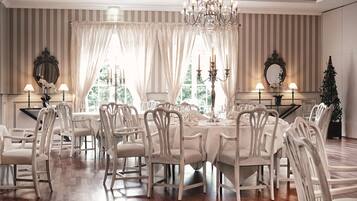 A dining room with a large table set for a meal, surrounded by white chairs, with chandeliers and wall-mounted mirrors.