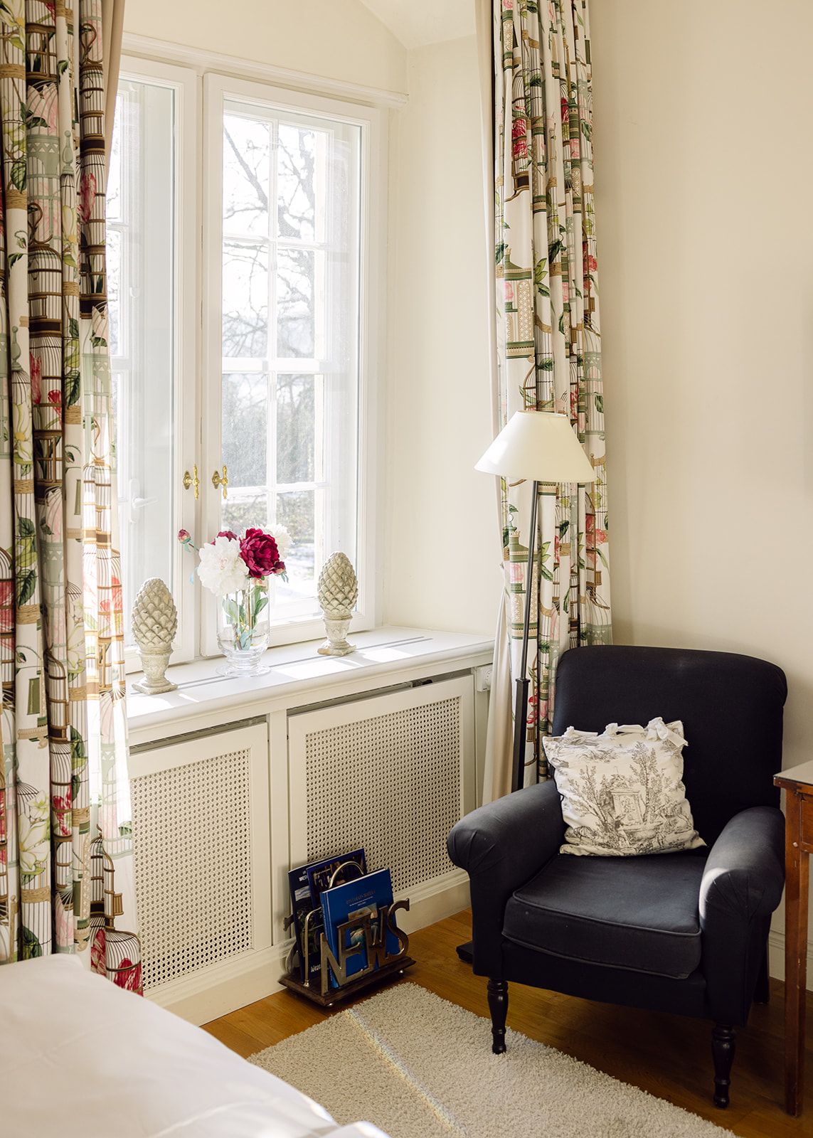 Una habitación con un sillón negro, una ventana con cortinas, una lámpara y un radiador.
