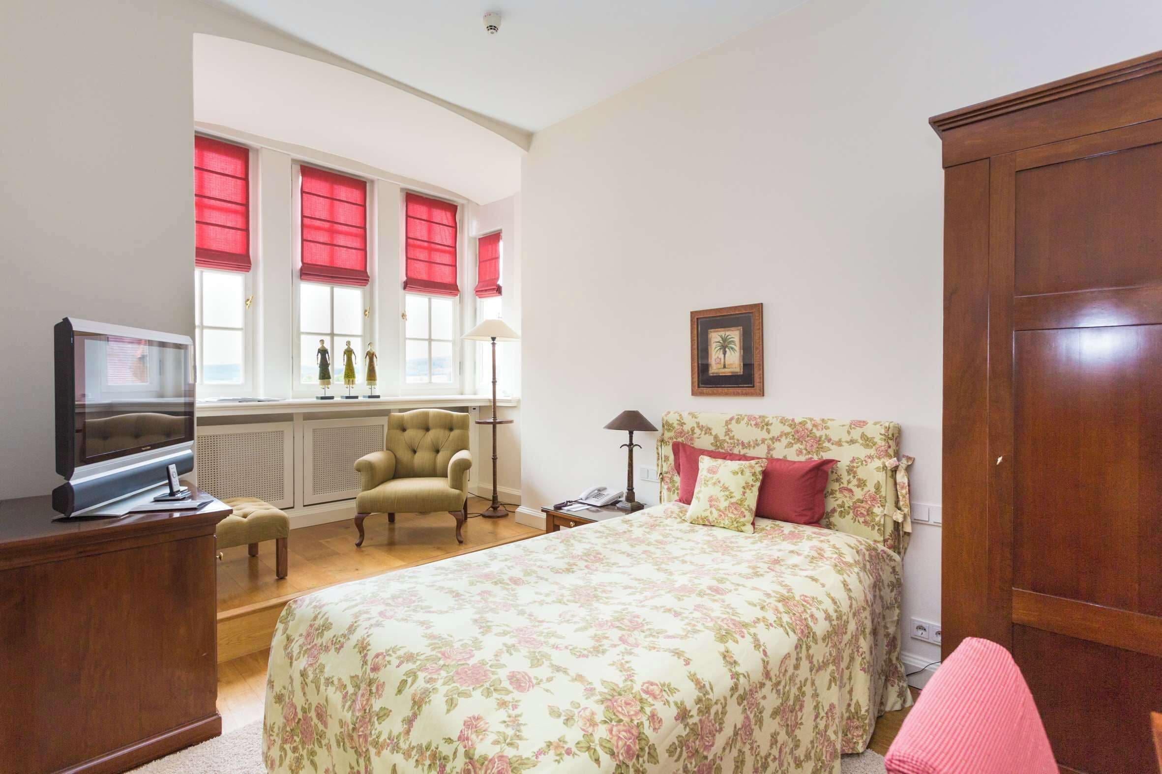 Ein Schlafzimmer mit Bett, Stuhl, Fernseher, Kleiderschrank und einem Fenster mit roten Jalousien.