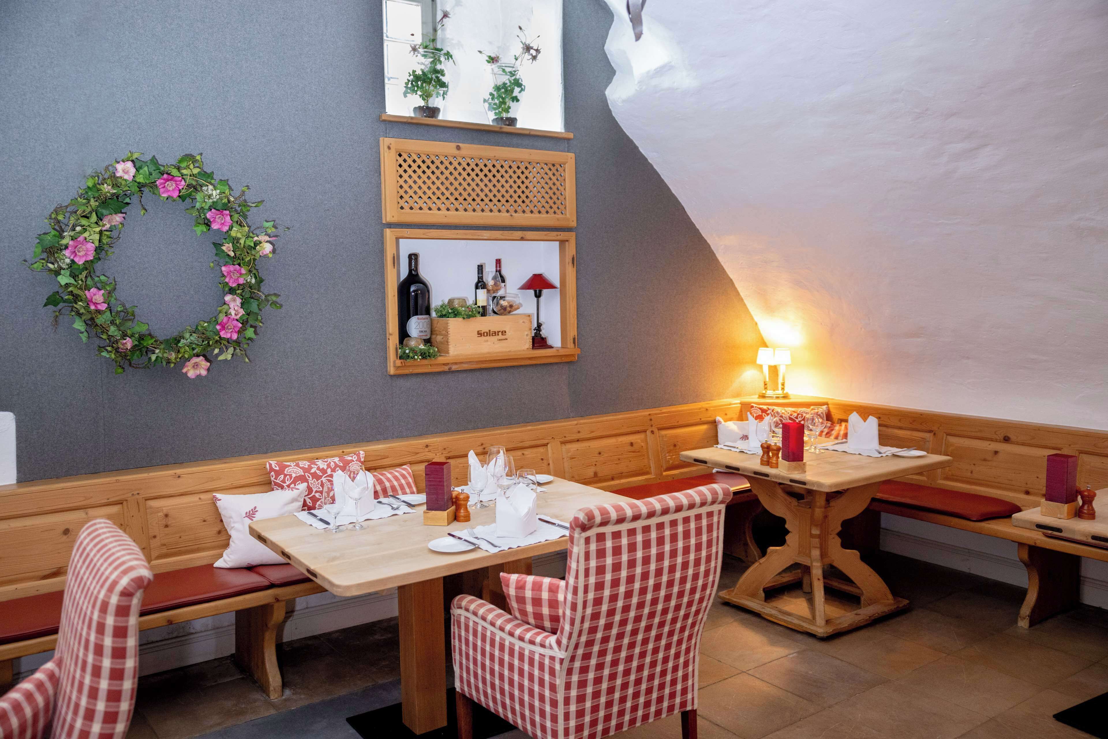 Een gezellige eethoek met houten banken en tafels, een bloemenkrans aan de muur en een plank met flessen en glazen.