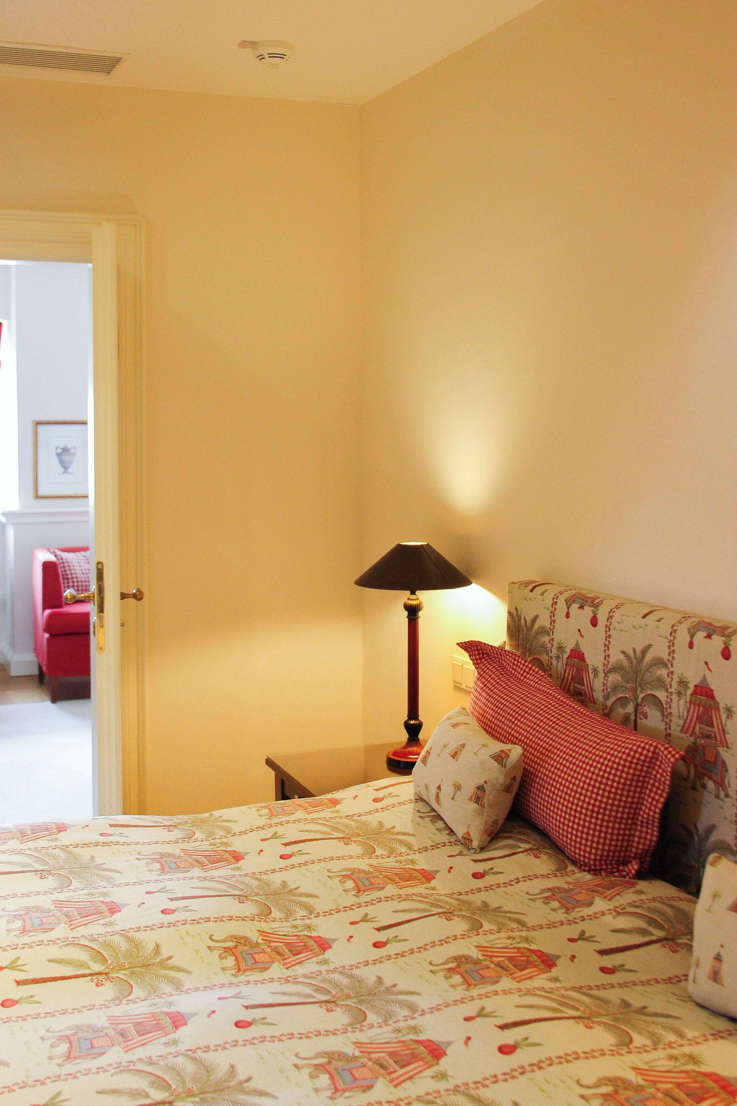A hotel room with a bed featuring a tropical print bedspread, a nightstand with a lamp, and a red chair in the background.