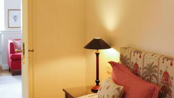 Habitación de hotel con una cama y una colcha con estampado tropical, una mesita de noche con lámpara y una silla roja al fondo.